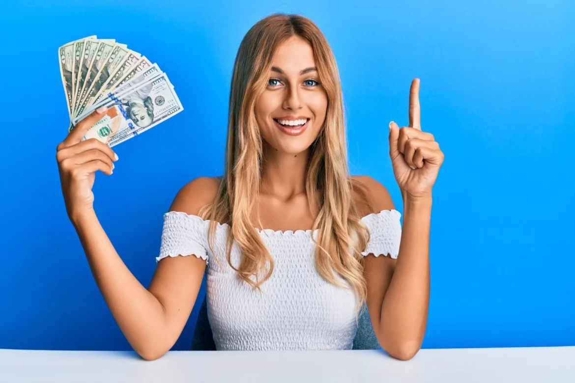 Beautiful blonde young woman holding dollars smiling with an idea or question pointing finger with happy face, number one