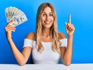 Beautiful blonde young woman holding dollars smiling with an idea or question pointing finger with happy face, number one